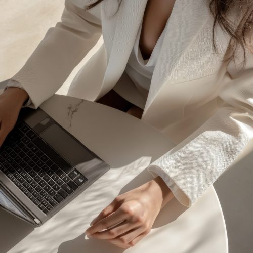 A woman in a white blazer sits at a round table with a laptop and a cup of coffee. Sunlight streams in, highlighting her focused expression as she engages with her work.
